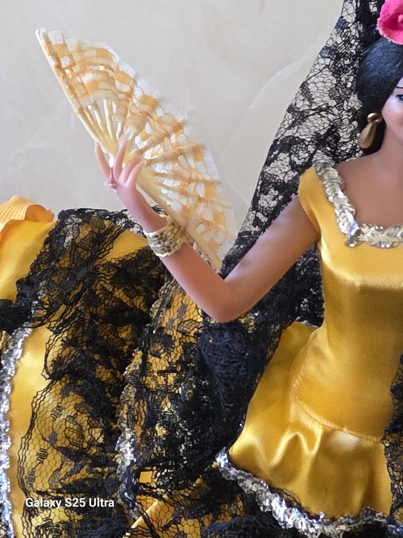 以下が含まれることがあります： 黒髪にピンクの花、金の装飾を施した、黒いレースのトリムが付いた黄色のドレスを着た人形。ドレスは首と袖に銀色のトリムが付いています。人形は扇子を持っており、扇子は黄色と白です。