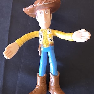 May include: A Woody action figure from the Toy Story franchise. The figure is wearing a brown cowboy hat, yellow shirt, blue pants, and brown boots. The figure has its arms outstretched.
