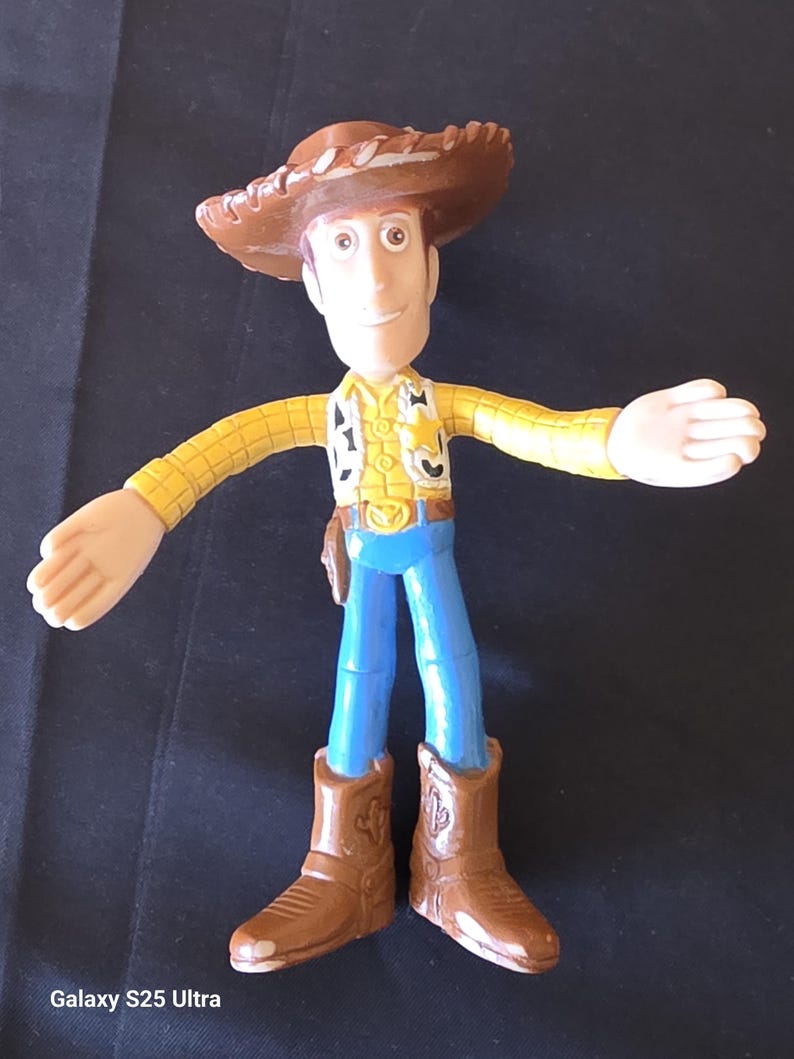 May include: A Woody action figure from the Toy Story franchise. The figure is wearing a brown cowboy hat, yellow shirt, blue pants, and brown boots. The figure has its arms outstretched.