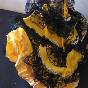 以下が含まれることがあります： 黄色い衣装で、黒いレースのトリムとアクセントが付いています。 レースは複雑な模様で、黄色の生地の上に重ねられています。 衣装にはフリルの縁があり、コスチュームまたはフォーマルウェアの一部のように見えます。