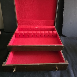 May include: A dark brown wooden storage chest with a hinged lid and a single pull-out drawer. The interior is lined with bright red felt. The upper section features a slotted insert for organizing flatware or utensils, while the drawer is open.