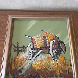 May include: A framed painting depicts a rustic wooden wagon in a field. The wagon is painted in shades of brown and yellow, with white wheels. The background is green with a fence and tall grass. The painting is signed "Abbrook".