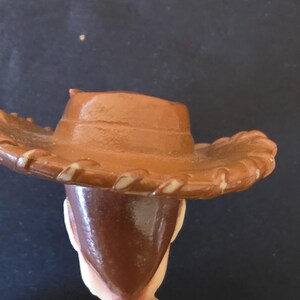 May include: A close-up of a toy figure of a cowboy, wearing a brown hat with a light brown trim. The figure has brown hair and a yellow shirt with a yellow neckerchief. The background is dark.