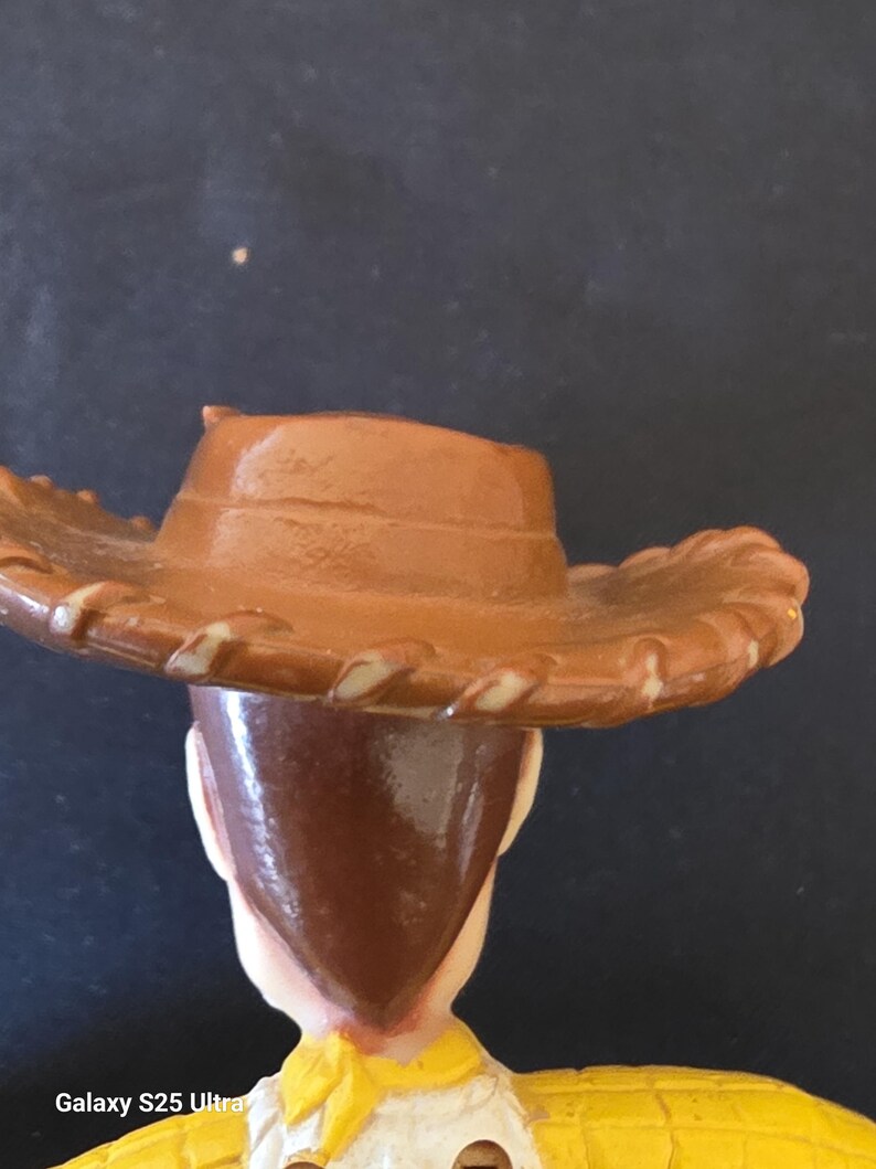 May include: A close-up of a toy figure of a cowboy, wearing a brown hat with a light brown trim. The figure has brown hair and a yellow shirt with a yellow neckerchief. The background is dark.