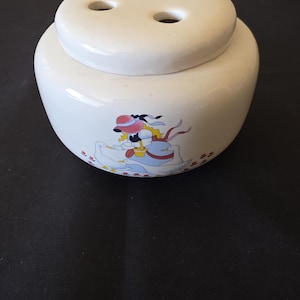 May include: A white ceramic flower frog or posy vase with a rounded body and a removable lid featuring four circular openings. The side of the vase is decorated with a colourful illustration of a figure in a pink hat surrounded by three white geese.