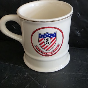 May include: A white ceramic mug with a gold-toned rim and a handle on the left side. The front features a circular emblem with a red rope border, a shield with blue stars, red and white stripes, and the text U.S.S. RANGER CV-61 in red letters.