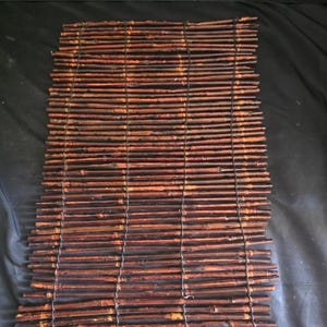 May include: A rectangular placemat constructed from numerous thin, dark brown bamboo sticks arranged horizontally and secured together with thin black cord. The natural texture of the wood is visible throughout the individual bamboo pieces.