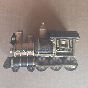 Peut inclure: Figurine décorative en forme de train, dans les tons marron, noir et or. Le train présente un design détaillé avec un conducteur visible dans la cabine. Les roues et autres accents sont mis en valeur par des bordures dorées.