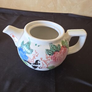May include: White ceramic teapot with a painted floral and bird design. The teapot features blue, pink, and green flowers, along with a black and white bird. The handle and spout are white, and the overall design is detailed and colorful.