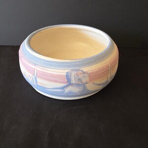 May include: A round ceramic bowl with a matte, textured exterior featuring horizontal bands of pastel pink, light blue, and cream. The interior of the bowl is a smooth, off-white color. The design includes stylized, abstract blue shapes.