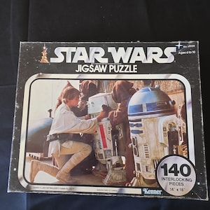 May include: A Star Wars jigsaw puzzle box featuring a scene of Luke Skywalker interacting with R2-D2 and a Jawa. The black box displays the Star Wars logo, 140 interlocking pieces, and dimensions of 35.56 cm by 45.72 cm, with Kenner branding.