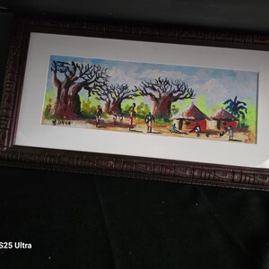 May include: A framed painting depicting a landscape with large baobab trees, small huts with thatched roofs, and several figures in an open field. The artwork features a light blue sky, green foliage, and is signed M. UNGA in the lower left corner.