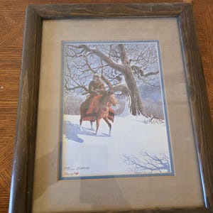 May include: Framed print depicting a person on horseback in a snowy landscape. The artwork features a rider in winter attire, a brown horse, and a bare tree. The title "THE INTRUDER" is on a small plaque.