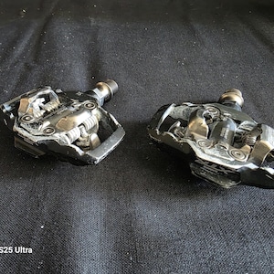 May include: A pair of black and silver Shimano XTR clipless bicycle pedals featuring a metallic body, spring-loaded retention mechanisms, and threaded spindles. The pedals show signs of wear on the outer edges and metallic surfaces.