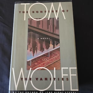 Puede incluir: Un libro de tapa dura titulado "La hoguera de las vanidades" de Tom Wolfe. La portada presenta un paisaje urbano reflejado en una ventana. El nombre del autor se muestra de forma destacada. Es una novela.