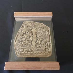 May include: A beige-colored, rectangular relief sculpture depicting a detailed scene with figures and a tree, encased in a clear acrylic display with a wooden frame. The sculpture is likely of historical or cultural significance.