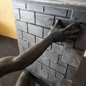 K&ouml;nnte beinhalten: Eine dunkelgraue Backsteinmauer mit dekorativer Oberseite. Ein dunkelgrauer Arm und eine Hand greifen nach der Mauer. Die Ziegel haben ein strukturiertes Aussehen.