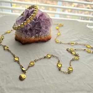 Op de afbeelding: Een goudkleurige ketting en armband set met gele edelsteen accenten. De ketting heeft een delicate ketting met gelijkmatig verdeelde stenen, en de armband heeft een bijpassend ontwerp. De sieraden worden gepresenteerd op een paars-oranje kristal.