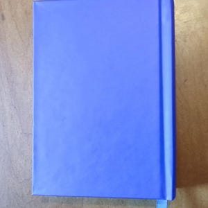 May include: A closed, rectangular notebook with a solid blue cover. A matching blue ribbon bookmark extends from the top edge. The notebook is set against a light brown wooden surface.