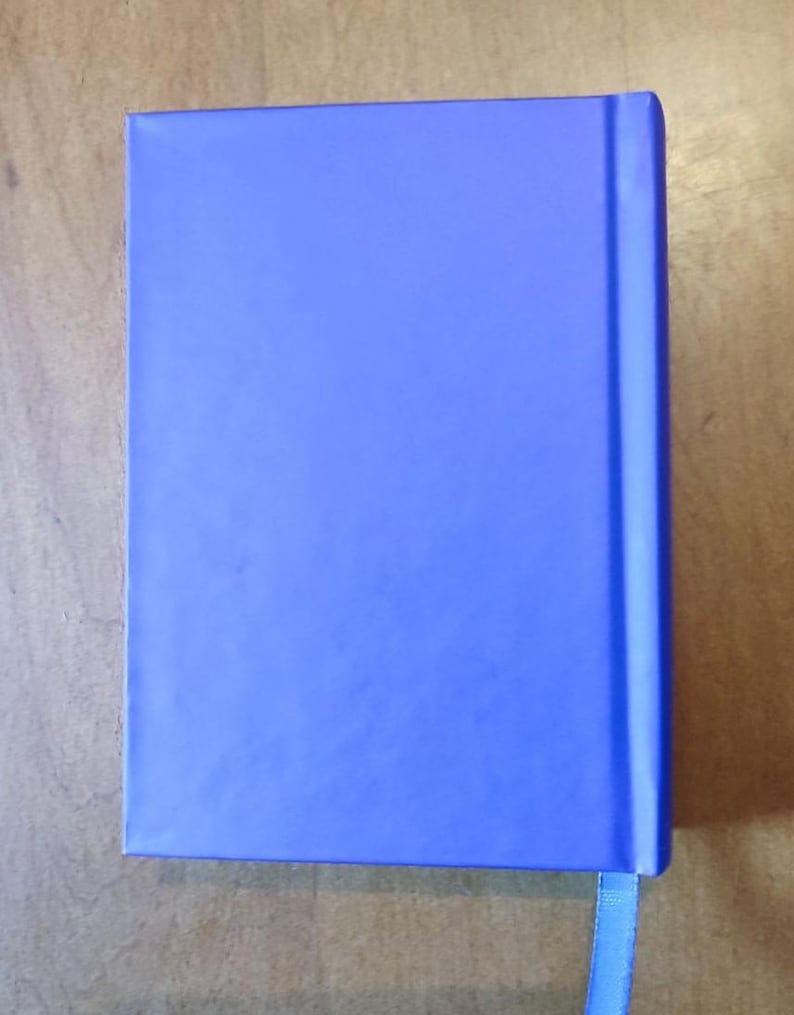 May include: A closed, rectangular notebook with a solid blue cover. A matching blue ribbon bookmark extends from the top edge. The notebook is set against a light brown wooden surface.