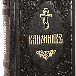May include: Black leather-bound book with gold lettering and a cross emblem. The cover features ornate embossed designs and the word "KANONNIKZ" in gold. The spine also has gold lettering. A religious item.