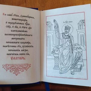 May include: An open book with blue covers, featuring text and an illustration. The left page displays Cyrillic script in black and red ink, while the right page shows a line drawing of a crowned figure playing a harp, framed in red.