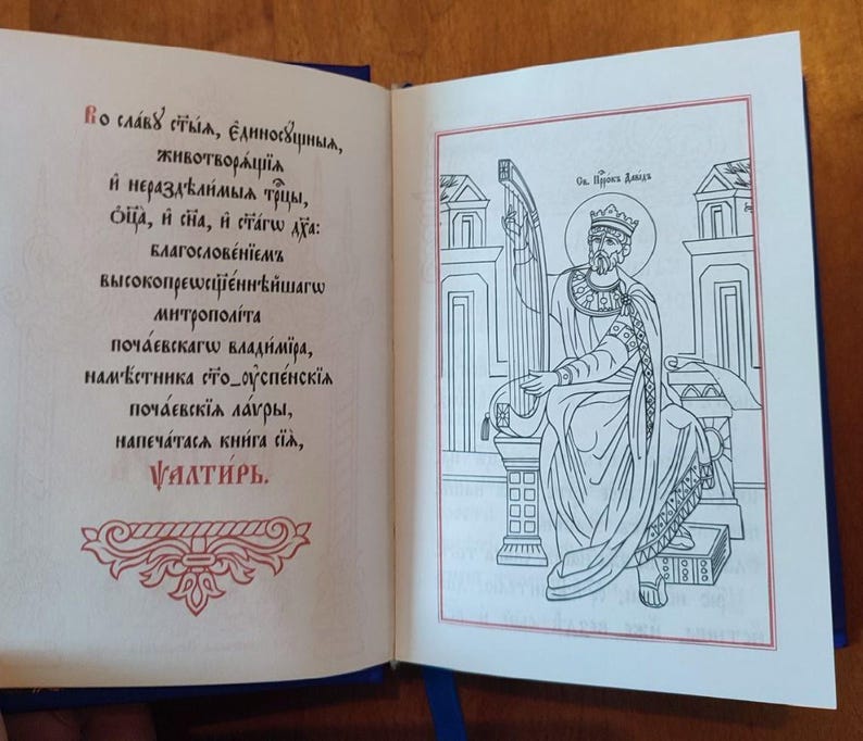 May include: An open book with blue covers, featuring text and an illustration. The left page displays Cyrillic script in black and red ink, while the right page shows a line drawing of a crowned figure playing a harp, framed in red.