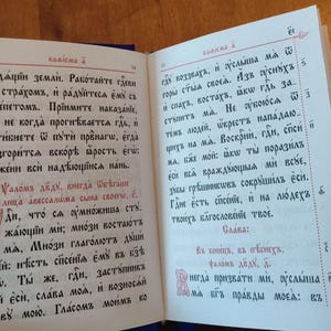 May include: An open book with text in an old Slavic script. The pages are cream-colored with red and black ink. The text is arranged in columns with decorative initial letters.