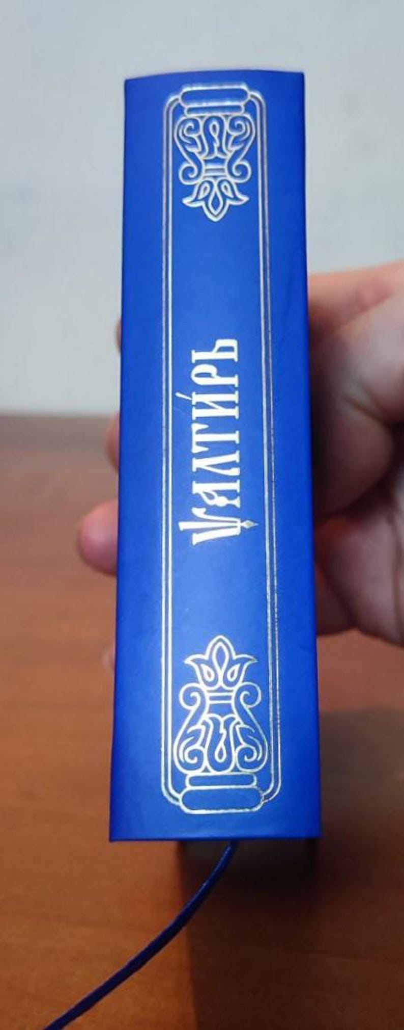 May include: A blue book spine with ornate silver-colored designs and text. The text reads "Уалтирь" in Cyrillic script. A blue bookmark is attached.