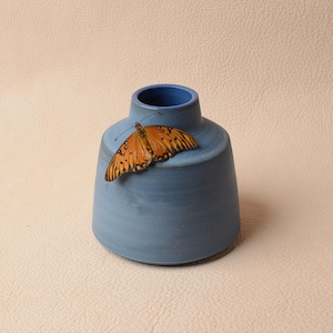 May include: A small, blue ceramic vase with a flared base and a narrow neck. An orange butterfly with black markings rests on the rim of the vase. The background is a neutral, textured surface.