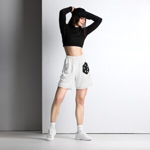 May include: White athletic shorts with a black pocket and a black design. The shorts have a drawstring waist and a black design on the left leg. The model is wearing a black cap and a black long-sleeve shirt.