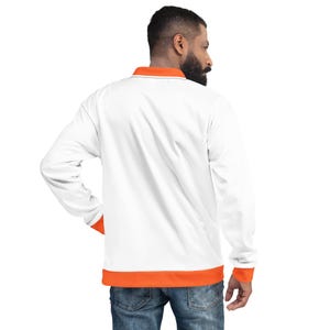 Puede incluir: Chaqueta blanca de manga larga con ribete naranja en el cuello, los puños y el dobladillo. La chaqueta tiene cuello y un diseño sencillo. El modelo lleva vaqueros azules.