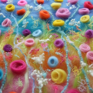 A close up photo of the fibres laid out before they are wet felted. The blue, yellow and pink background fibres are scattered with yarn for stems, and scraps of colourful scrim, wool nepps and twirls of merino to make an abundance of meadow flowers