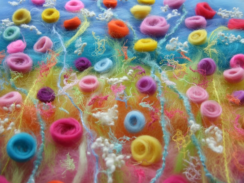 A close up photo of the fibres laid out before they are wet felted. The blue, yellow and pink background fibres are scattered with yarn for stems, and scraps of colourful scrim, wool nepps and twirls of merino to make an abundance of meadow flowers