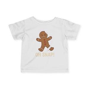 May include: White t-shirt featuring a distressed gingerbread man graphic with a bite taken out of its head and leg. The text "OH SNAP!" is printed below the gingerbread man in a tan color.