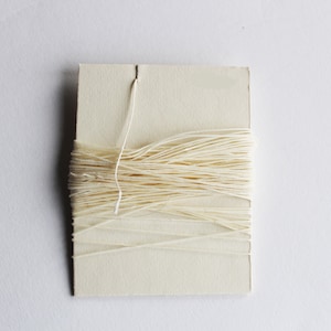 May include: A card of off-white thread, neatly wound and secured. A single strand of the thread emerges from a slit at the top of the card. The thread is tightly packed, creating a tidy, organized look.