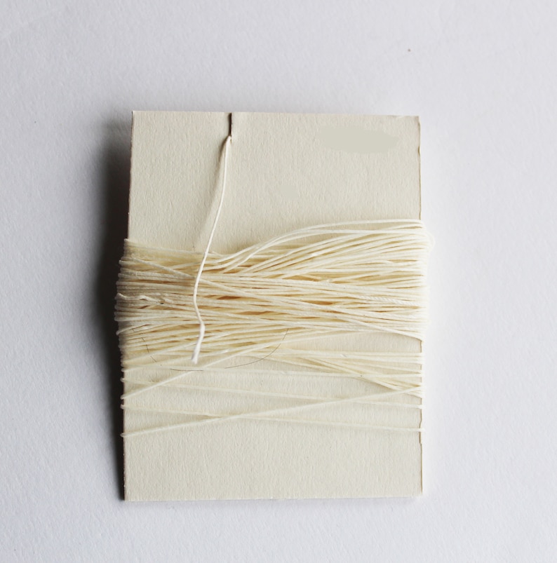 May include: A spool of off-white thread wrapped around a rectangular cardboard card. A single strand of thread extends from a slit at the top of the card. The thread is tightly wound, creating a neat, organized appearance.