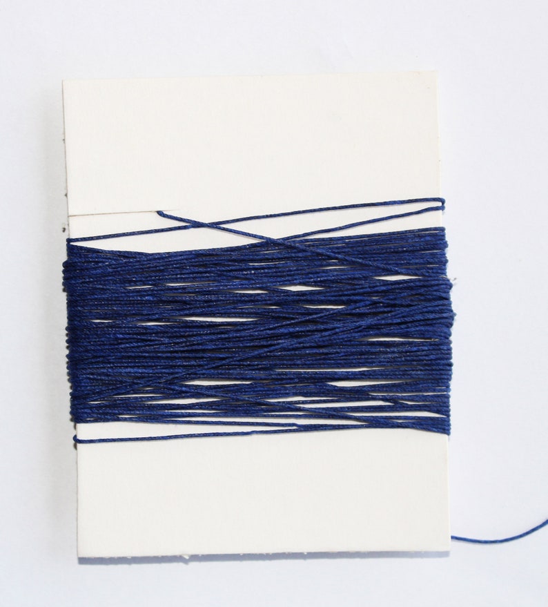 May include: A spool of navy blue thread is wrapped around a white rectangular card. The thread is tightly wound, creating a dense, textured appearance. A single strand of thread extends from the spool, suggesting it's ready for use in sewing or crafting projects.