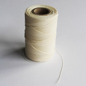 May include: A cylindrical spool of cream-coloured thread, tightly wound around a cardboard core. The thread is smooth and appears to be made of a natural fibre. A length of thread extends from the spool. Suitable for sewing, crafting, or textile projects.