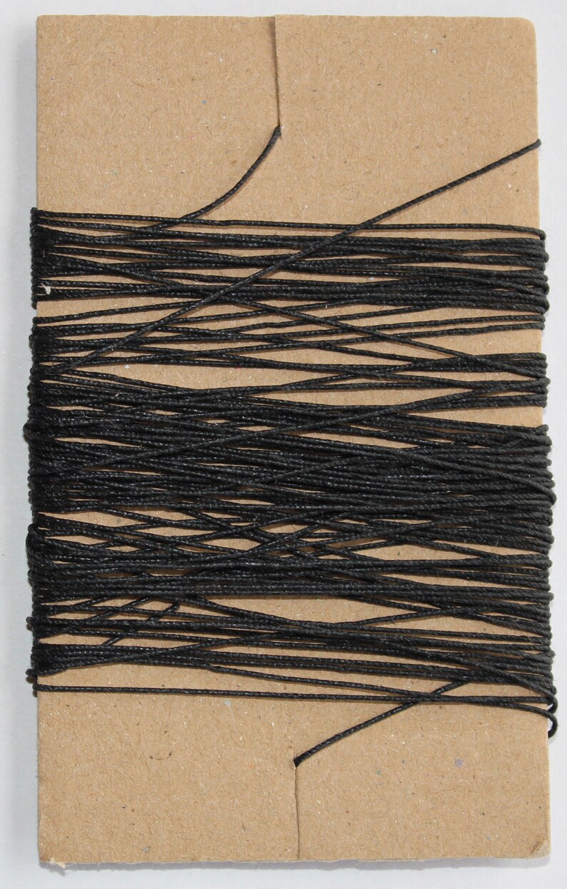 May include: A spool of black thread wrapped around a rectangular cardboard card. The thread is tightly wound, creating a dense, dark mass. The card is a light brown color.