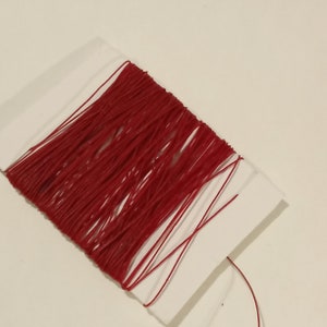 May include: A spool of crimson thread, tightly wound on a white card. The thread has a rich, saturated colour and a slight sheen. The image is a close-up, highlighting the texture and colour of the thread, suitable for sewing or crafts.