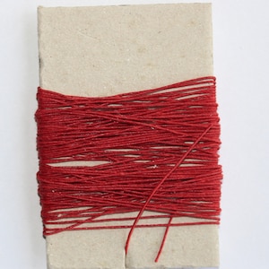 May include: A rectangular cardboard spool wrapped with bright red thread. The thread is tightly wound around the cardboard, with a few loose strands extending from the main bundle. The background is plain white.