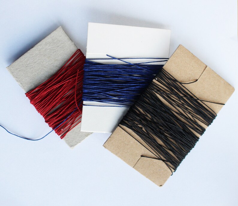 May include: Three spools of thread in red, blue, and black. The thread is wound around cardboard spools. The spools are arranged diagonally on a white surface. The thread appears to be a fine, smooth material.