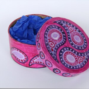 May include: A round, open pink box with a lid, decorated with a paisley design in shades of pink, purple, and red. The box contains blue tissue paper. The lid is partially open, revealing the contents.