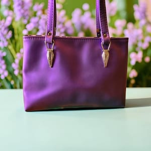 May include: A rectangular purple handbag with a smooth, slightly iridescent finish. The bag features two vertical straps attached to the top edge with silver-toned, triangular metal hardware connectors that provide a secure and structured look.
