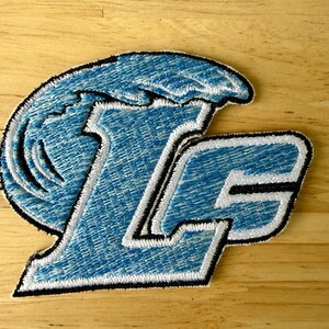 Könnte beinhalten: Ein gestickter Aufnäher mit den Buchstaben LG in einer fetten Blockschrift. Die Buchstaben sind mit hellblauem Garn gefüllt und mit Weiß und Schwarz umrandet. Eine stilisierte hellblaue Wellengrafik wölbt sich über den Buchstaben L.