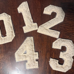 May include: Five off-white felt numbers: 0, 1, 2, 3, and 4. The numbers have a textured, fuzzy appearance and are arranged on a dark brown wooden surface. The numbers are likely for crafts or personalization.