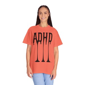 Op de afbeelding: Een koraalkleurig T-shirt met het woord "ADHD" in zwarte, druipende letters. Het shirt heeft korte mouwen en een ronde hals. Het model draagt een blauwe spijkerbroek.