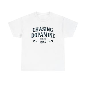 May include: White t-shirt featuring the dark blue text "CHASING DOPAMINE since 1982". The text is centrally positioned. The shirt has a round neck and short sleeves.