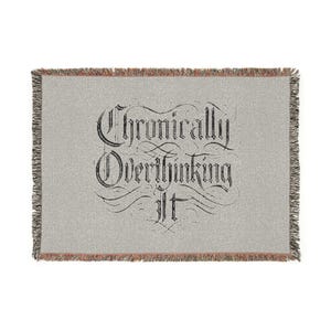 Peut inclure: Une couverture grise claire avec l'inscription "Chronically Overthinking It" en noir. La couverture a une bordure à franges avec un mélange de gris, de marron et d'orange.